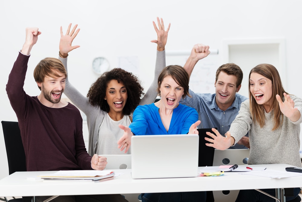 Excited successful business team of diverse multiethnic young people sitting at a table in the office cheering exuberantly as they celebrate a successful outcome on the laptop computer